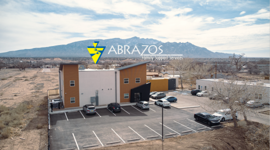 Abrazos logo superimposed on a photo of the Abrazos campus with a 2-story building and two 1-story buildings against the backdrop of the Sandia mountains